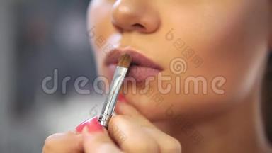 在模特的嘴唇上涂上专业的化妆，用金色光泽`画笔的特写。 嘴唇上的光泽
